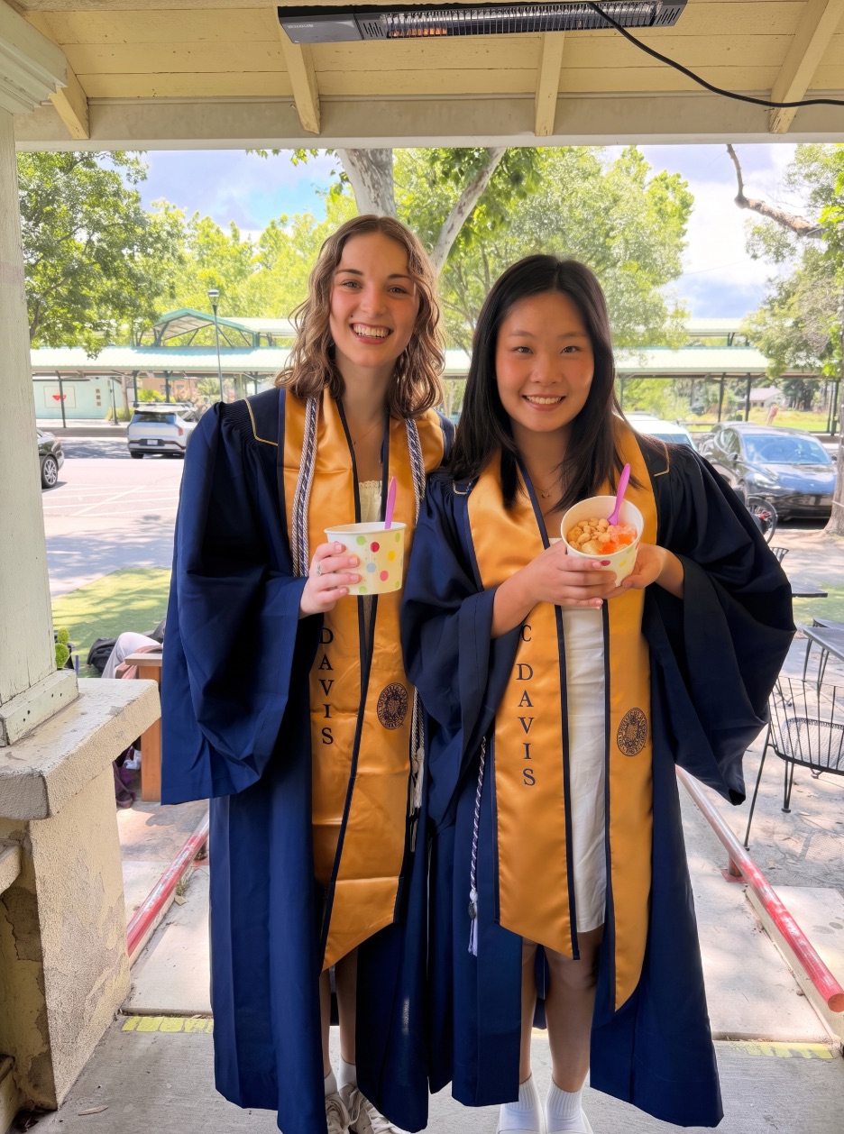 Two UC Davis graduates in caps and gowns holding cups of frozen yogurt